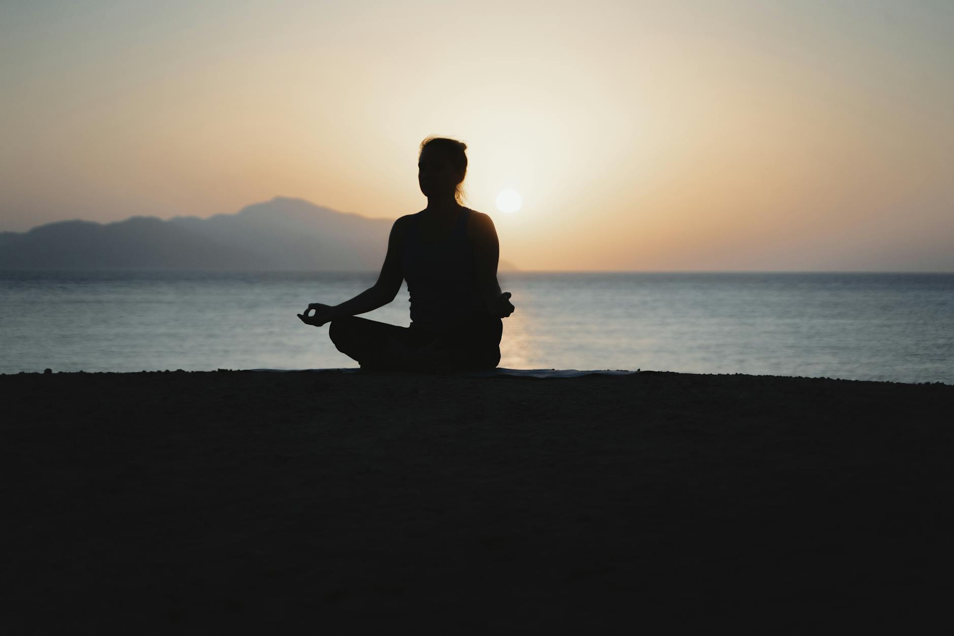 Person sitting cross-legged and meditating at sunset by the ocean.
